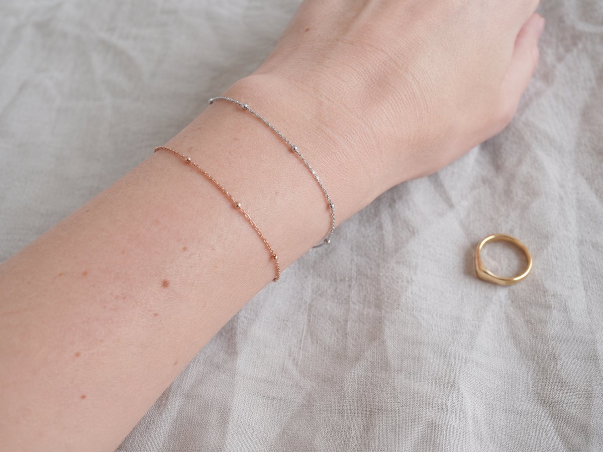 Poignet portant deux bracelets fins argent et or rose, avec une petite bague dorée posée sur un tissu beige