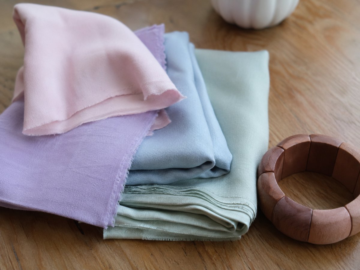 Carrés de tissu en tons pastel rose, lilas, bleu ciel et vert sauge posés sur une table en bois avec un bracelet en bois