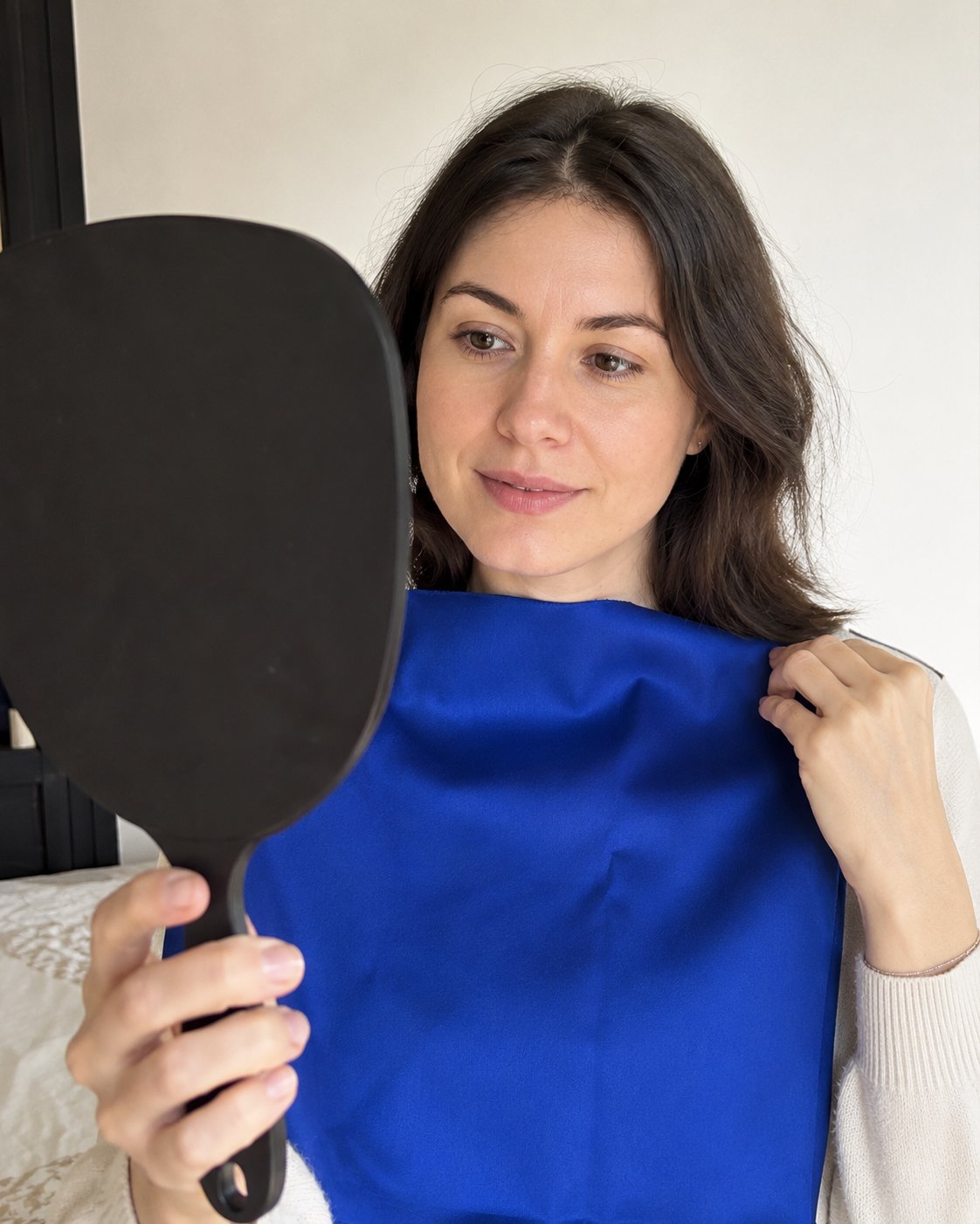 Femme tenant un tissu bleu royal près du visage et se regardant dans un miroir à main, test de draping colorímétrique